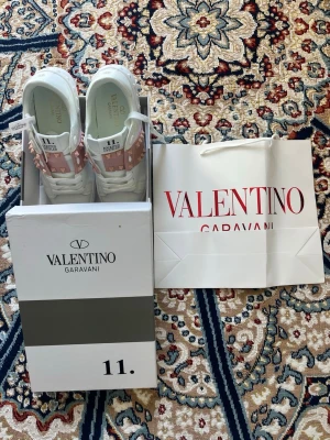 Valentino Rockstud Untitled sneakers - Snygga vita sneakers från Valentino Garavani med rosa band och ikoniska nitar över vristen. Modellen heter Rockstud Untitled och har en clean siluett med exklusiv känsla. Skorna är tillverkade i läder och har vita skosnören. Perfekta för dig som vill sticka ut.