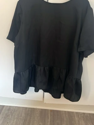 Svart satin blus med volangkant - Svart satin blus med volangkant nedtill. Blusen har rund halsringning och korta ärmar.