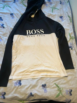 Svartvit hoodie från Hugo Boss - Snygg hoodie från Hugo Boss i svart och vitt med stor BOSS-logga på bröstet. Tröjan har huva och långa ärmar, perfekt för en avslappnad och trendig stil. Tillverkad i mjukt material som känns skönt mot huden.