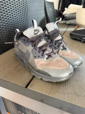 Nike Air Max 90 drift grå sneakers - Säljer ett par Nike Air Max 90 sneakers i olika grå nyanser med rutmönstrad mesh på tån och klassisk synlig Air-sula. Skorna har mörkgrå snören, detaljer i läder och textil samt den ikoniska Swoosh-loggan på sidan. Perfekta för dig som gillar streetwear och sportig stil.