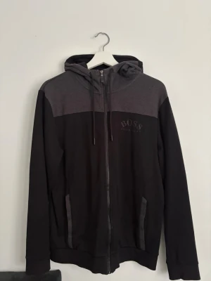 Svart och grå hoodie från BOSS - Snygg hoodie från BOSS i svart och mörkgrått med dragkedja framtill och två fickor. Huva med dragsko och diskret BOSS-logga på bröstet. Tillverkad i mjukt bomullsmaterial som känns skönt mot huden. Perfekt för en avslappnad och stilren look.