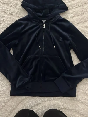 Mörkblå velour hoodie Juicy Couture - Snygg mörkblå hoodie från Juicy Couture i mjuk velour. Dragkedja framtill, fickor och justerbar huva med snören. Diskret Juicy-logga på bröstet. Perfekt för en chill och trendig look.
