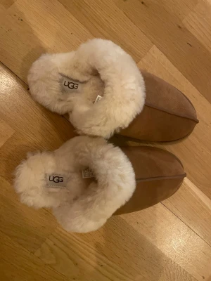 Oanvända UGG original- tofflor med fårskinn - Mysiga bruna tofflor från UGG med fluffigt vitt fårskinnsfoder. Tofflorna har en klassisk slip-in modell och är tillverkade i mocka på utsidan. Perfekta för att hålla fötterna varma hemma under kalla dagar. Tofflorna är oanvända. 
