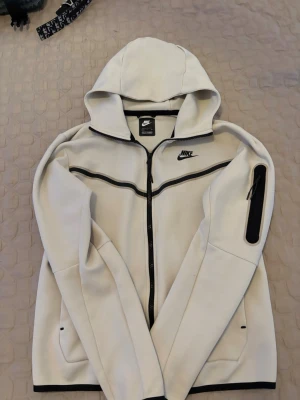 Nike tech  - Snygg vit hoodie från Nike med hel dragkedja och svarta detaljer. Hoodien har en huva, fickor på sidorna och en extra ficka med dragkedja på ärmen. Tillverkad i mjukt och bekvämt material, perfekt för en sportig och avslappnad stil.