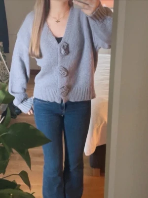 Ljuslila/grå stickad kofta med rosetter - Säljer en söt ljuslila/gråblå stickad kofta med tre dekorativa rosetter framtill. Koftan har v-ringning, långa ärmar och ribbade muddar vid ärmslut och nederkant. Perfekt för dig som gillar en gullig och trendig stil. Bara använd fåtal gånger och inte defekter!!