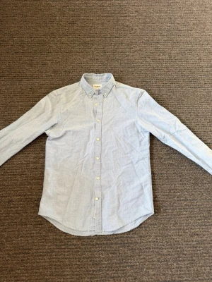 Ljusblå skjorta - Klassisk ljusblå från pull and bear med button-down krage och vita knappar. Skjortan har lång ärm och rundad nederkant. Tillverkad i ett mjukt bomullstyg som ger en avslappnad men stilren look.