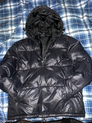 Svart pufferjacka Zara - Snygg svart pufferjacka från Zara med dragkedja och stor luva. Jackan har en glansig yta och breda quiltade partier som ger en modern look. Perfekt för kalla vinterdagar och enkel att matcha med olika outfits.