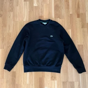 Svart sweatshirt från Lacoste - Klassisk svart sweatshirt från Lacoste med rund halsringning och den ikoniska krokodilloggan broderad på bröstet. Tröjan har ribbade muddar vid ärmslut och nederkant samt en relaxed classic fit. Perfekt för en clean och stilren look. Pris kan diskuteras, skriv gärna bud. Nypris 1595kr. 