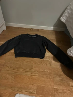Svart croppad sweatshirt - En svart croppad sweatshirt med rund hals och långa ärmar. Tröjan har en enkel och clean design utan tryck eller detaljer. Perfekt till jeans eller mjukisbyxor för en chill look. Materialet ser ut att vara mjukt och bekvämt, troligen bomull eller bomullsblandning.