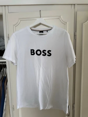 Vit BOSS t-shirt med tryck - Säljer en klassisk vit t-shirt från BOSS med svart BOSS-tryck på bröstet. T-shirten har rund halsringning och korta ärmar. Tillverkad i mjuk bomull som känns skön mot huden. Perfekt till jeans eller shorts för en clean och enkel stil.