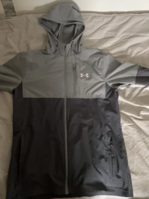 Grå vindjacka från Under Armour - Snygg tvåfärgad vindjacka från Under Armour i ljusgrått och mörkgrått. Jackan har huva, hel dragkedja framtill, bröstficka med dragkedja och logga på bröstet. Tillverkad i lätt och tåligt material som står emot vind.