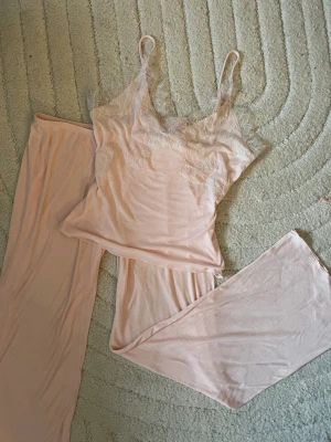Ljusrosa spets-set med linne och byxa - Säljer ett ljusrosa pyjamas set med linne och långa byxor. Linnet har tunna axelband och v-ringning dekorerad med mjuk spets. Materialet är tunt och stretchigt, vilket gör det bekvämt och luftigt. Perfekt för dig som gillar en romantisk och feminin stil.