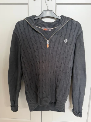 Mörkgrå stickad tröja från Morris - Mörkgrå kabelstickad tröja från Morris med halv dragkedja och ribbade muddar. Tröjan har en liten vit logotyp på bröstet och en bred krage som kan vikas ner. Perfekt för kyliga dagar och snygg till jeans.