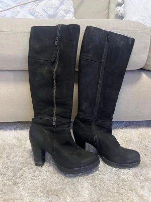 Svarta höga stövlar med klack - Snygga svarta stövlar i mockaimitation med grov klack och dragkedja längs sidan. Stövlarna är knähöga och har en stilren design som passar till många outfits. Perfekta för dig som vill ha både höjd och edge i din look.