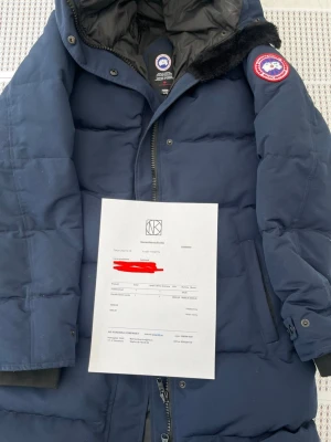 Mörkblå dunjacka från Canada Goose - Snygg mörkblå dunjacka från Canada Goose med klassisk patch på ärmen och huva med svart fuskpäls. Jackan har dragkedja, tryckknappar och ribbade muddar vid ärmsluten. Perfekt för kalla vinterdagar och riktigt varm.