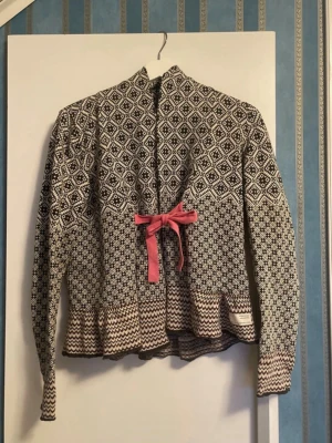 Mönstrad stickad kofta med rosett - Unik stickad kofta med svartvitt geometriskt mönster och beige detaljer. Koftan har en rosa rosett framtill som knyts i midjan och vågig kant nedtill. Perfekt för dig som gillar retrovibbar och snygga detaljer.