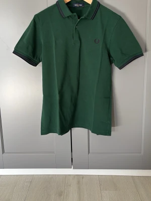 Grön pikétröja från Fred Perry - Klassisk mörkgrön pikétröja från Fred Perry med svart broderad logga på bröstet. Tröjan har krage, knappar vid halsen och svarta detaljer på ärmslut och krage. Perfekt för en stilren och avslappnad look.