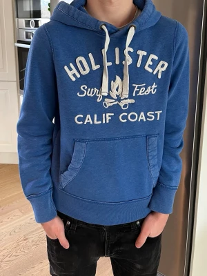 Blå Hollister hoodie med tryck - Snygg blå hoodie från Hollister med vit text. Hoodien har en huva med vita dragsnören och en klassisk magficka. Perfekt för chill dagar och streetwear-stil. 