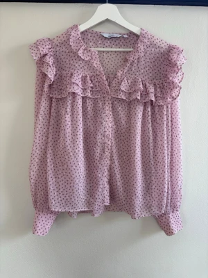 Rosa blommig volangblus från Lindex - Supersöt rosa blus från Lindex med små blommor, volangdetaljer över bröstet och axlarna samt knappar framtill. Blusen är luftig och har lång ärm med bred manschett. Perfekt för dig som gillar romantisk och feminin stil.