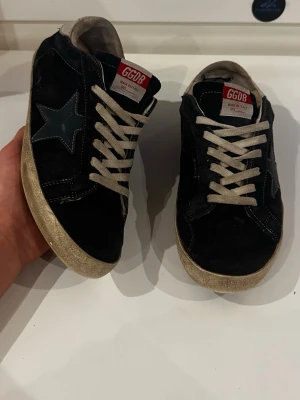 Svarta GGDB sneakers - Snygga svarta sneakers från Golden Goose med vit sula och vita skosnören. Skorna har en ikonisk stjärndetalj på sidan och GGDB-logga på plösen. Ovandelen är i mocka och läder, vilket ger en cool och avslappnad vibe.