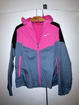Nike vindjacka rosa och grå - Snygg vindjacka från Nike i rosa och grått med svarta detaljer. Jackan har huva, dragkedjeförsedda fickor och en sportig design. Perfekt för blåsiga dagar och outdoor-aktiviteter. Tillverkad i lätt och tåligt syntetmaterial.