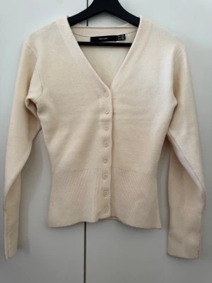 Beige ribbad kofta från Vero Moda - Beige tröja med V-ringning och långa ärmar. Koftan har ribbad midja och har matchande färg knappar framtill. Tillverkad i mjukt stickat material som ger en jätte skön passform, till och med ganska varm, jag gillar inte hur tajt den sitter på mig därmed är denna ALDRIG använd asså alls. Den har bara hängt i mitt skåp 🥱 