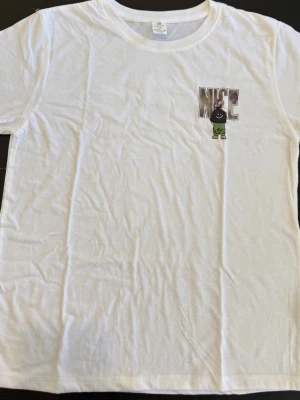 Vit t-shirt med NICE-tryck - Vit T-shirt med “NICE”-tryck – streetwear (M/L)  Snygg vit T-shirt med tryck av texten “NICE” och en tecknad figur med lila keps och gröna byxor på både fram- och baksida. Stort och iögonfallande tryck baktill. Klassisk rund hals och korta ärmar ger en bekväm passform. Perfekt för en avslappnad och trendig streetwear-look.  Storlek: M–L  💬 Hör av dig vid intresse!