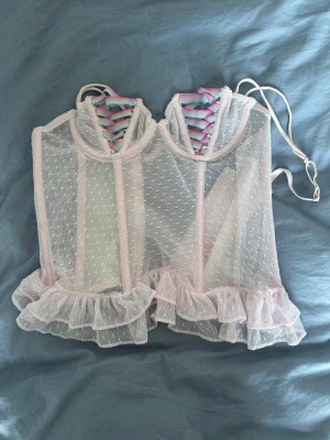 Rosa meshkorsett med volangkant - Söt korsett i ljusrosa mesh med små prickar och volangkant nedtill. Axelbanden är justerbara och framtill finns en dekorativ snörning i pastellfärger. Materialet är genomskinligt och ger en romantisk känsla.