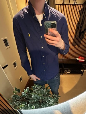 Mörkblå skjorta från Ralph Lauren - Klassisk mörkblå skjorta från Ralph Lauren med button-down krage och gul broderad logga på bröstet. Skjortan har långa ärmar, vita knappar och en slim passform. Perfekt till jeans för en clean och stilren look.