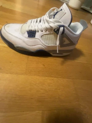 Vita Air Jordan 4 Retro sneakers - Snygga vita Air Jordan 4 Retro sneakers med blå detaljer och meshpaneler på sidorna. Skorna har en hög siluett, klassisk snörning och synlig Air-enhet i sulan. Tillverkade i läder och textil för en sportig och clean look.
