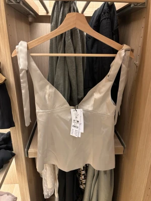 Champagnefärgad satinlinnetopp med knyt - Snygg champagnefärgad linnetopp i satin med breda axelband som knyts upptill. Toppen har v-ringning och en figurnära passform. Dragkedja bak för enkel påklädning. Perfekt för dig som vill ha en stilren och trendig look.