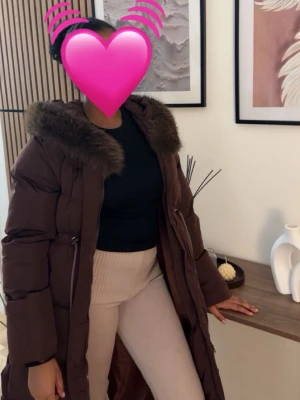 Brun lång pufferkappa med fuskpäls - Snygg brun lång pufferkappa med stor huva och fuskpälsdetalj runt kragen. Kappan har dragkedja, knytband i midjan och stora fickor på sidorna. Perfekt för kalla dagar och ger en trendig siluett.