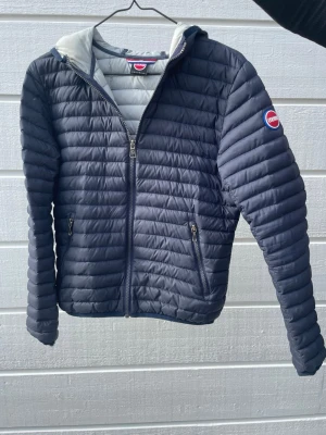 Mörkblå pufferjacka från Colmar - Snygg mörkblå pufferjacka från Colmar med quiltad design och huva. Jackan har dragkedja framtill, två fickor med dragkedja och en logotyp på ärmen. Insidan är ljusgrå och materialet är lätt och vadderat, perfekt för kalla vinterdagar.
