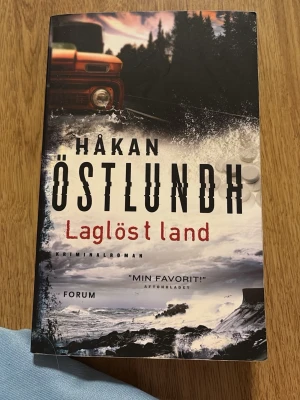 Laglöst land av Håkan Östlundh - Kriminalroman av Håkan Östlundh med titeln 'Laglöst land'. Omslaget har en dramatisk bild med en röd bil, mörka skogar och stormigt hav. Inuti finns en karta över Gotland. Perfekt för dig som gillar spännande deckare med svensk miljö. Sjätte delen av Östlundhs serie om Kriminalpolisen Fredrik Broman