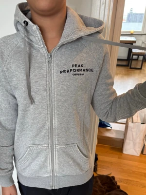 Grå hoodie Peak Performance - Snygg grå zip-hoodie från Peak Performance med svart logga på bröstet. Hoodien har dragkedja, två fickor fram och justerbar huva med snören. Tillverkad i mjukt sweatshirtmaterial som är skönt att ha på sig. Perfekt för en avslappnad stil.