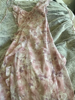 Blommig rosa maxiklänning med volanger - Säljer en romantisk maxiklänning i tunn, luftig mesh med rosa och lila blommönster. Klänningen har smala axelband och volangdetaljer längs kanterna. Perfekt för dig som gillar en feminin och somrig stil.