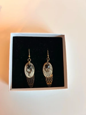 Unika örhängen i mässing och svart sten - Säljer ett par handgjorda örhängen i mässing med ovala hängen och snyggt ingraverat mönster. Örhängena har en svart pärlliknande sten i mitten som ger en cool kontrast till den guldiga metallen. Perfekta för dig som gillar unika accessoarer med vintagekänsla.