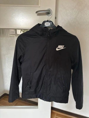 Svart vindjacka från Nike med huva - Snygg svart vindjacka från Nike med dragkedja framtill och två fickor med dragkedja. Jackan har huva och den klassiska Nike-loggan i vitt på bröstet. Tillverkad i lätt och tåligt material, perfekt för blåsiga dagar.