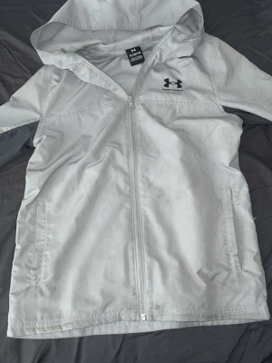 Ljusgrå vindjacka från Under Armour - Snygg ljusgrå vindjacka från Under Armour med dragkedja framtill, huva och två sidofickor. Jackan har en sportig look med diskret struktur i materialet och svart logga på bröstet. Perfekt för blåsiga dagar och enkel att matcha med andra plagg.