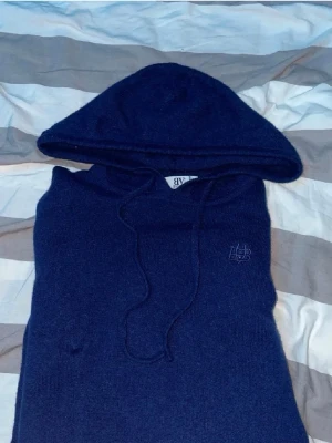 Mörkblå kashmire från Bvaldi - Mysig mörkblå hoodie från Bvaldi med broderad logga på bröstet. Tillverkad i mjukt stickat material och har justerbar dragsko i huvan. Perfekt för dig som gillar en enkel men stilren look.