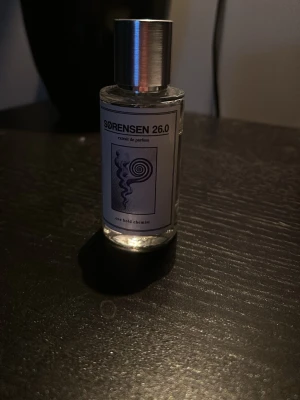 Sørensen 26.0 Extrait de Parfum - En stilren parfymflaska från Sørensen med metallock och transparent glas. Etiketten har en modern svartvit design med grafiskt mönster och texten 'Sørensen 26.0 extrait de parfum'. Flaskan är cylindrisk och minimalistisk.