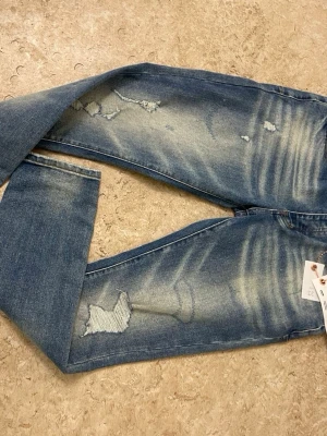 Blå slitna jeans med patchdetaljer - Säljer ett par blå jeans med slitningar och patchdetaljer på benen. Jeansen har en avslappnad passform och tydliga tvättade partier för en trendig look. Perfekta för dig som gillar en casual och edgy stil.