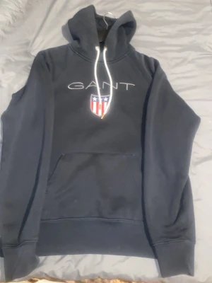 Mörkblå hoodie från GANT - Snygg mörkblå hoodie från GANT med vit dragsko i huvan och stor ficka framtill. På bröstet finns GANT-loggan och ett amerikanskt sköldmärke i rött, vitt och blått. Hoodien har ribbade muddar och är perfekt för en chill och sportig look. Strl M pris kan diskuteras 