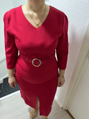 Röd midiklänning med bälte - Snygg röd midiklänning med trekvartsärmar och v-ringning. Klänningen har ett matchande bälte med rund metallspänne i midjan och en liten slits framtill. Dragkedja i ryggen för enkel påklädning. Materialet känns mjukt och följsamt. Är 164cm lång. Köpt utomlands och numret på storleken stämmer ej i Sverige