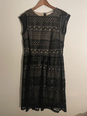 Svart spetsklänning med mönster - Säljer en svart spetsklänning med geometriskt mönster och beige underklänning. Klänningen har rund hals, är ärmlös och har en dragkedja i ryggen. Passformen är normal och längden går till knäna. Perfekt för dig som gillar unika detaljer och stilren design.