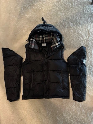 Burberry jacka/väst - Svart pufferjacka från Burberry med avtagbara ärmar och huva. Jackan har två dragkedjefickor framtill och ett klassiskt rutigt foder i grått, vitt och rött. Perfekt för kalla vinterdagar och kan bäras både som jacka och väst.