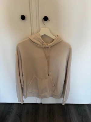 Beige hoodie med ficka framtill - Säljer en beige hoodie från BASICS i storlek S. Hoodien har en klassisk huva med dragsnören och en stor känguruficka framtill. Tillverkad i mjukt material som känns skönt mot huden. Perfekt för en avslappnad och stilren look.