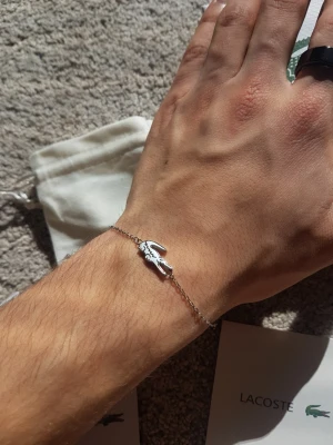 Silverfärgat armband från Lacoste - Snyggt och stilrent armband från Lacoste i silverfärgad metall. Armbandet har en tunn kedja och ett hängsmycke i form av den klassiska Lacoste-krokodilen. Perfekt accessoar för dig som gillar minimalistisk design och ikoniska detaljer.