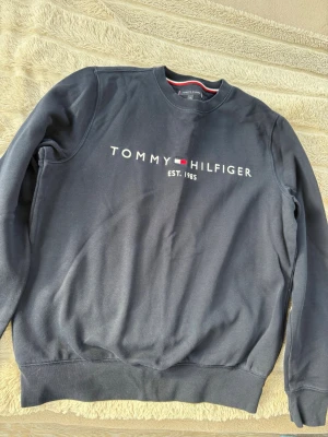Mörkblå sweatshirt Tommy Hilfiger - Snygg mörkblå sweatshirt från Tommy Hilfiger med rund halsringning och tryckt logga samt texten 'EST. 1985' på bröstet. Klassisk passform och mjukt material, perfekt till jeans eller joggers.