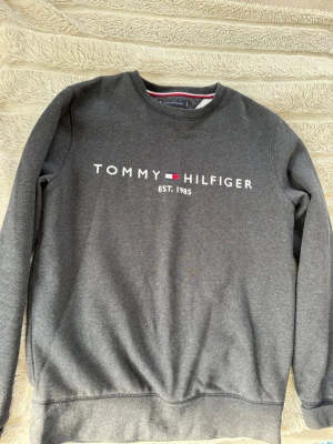 Mörkgrå sweatshirt Tommy Hilfiger - Mörkgrå sweatshirt från Tommy Hilfiger med klassisk logga och texten 'EST. 1985' på bröstet. Tröjan har rund halsringning, långa ärmar och mjukt material. Perfekt för en avslappnad och stilren look.